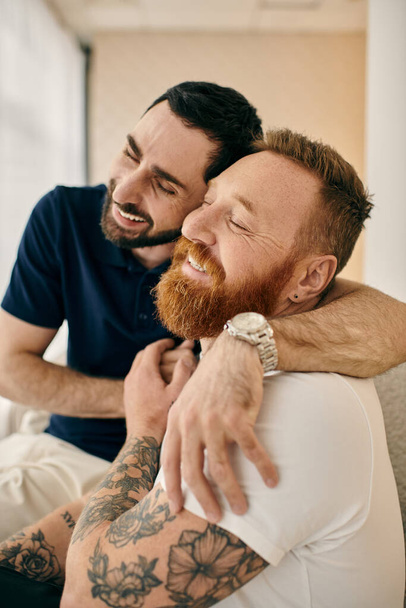 Zwei tätowierte Männer umarmen sich auf einer bequemen Couch in einem modernen Wohnzimmer liebevoll und spiegeln Liebe und Glück wider. - Foto, Bild