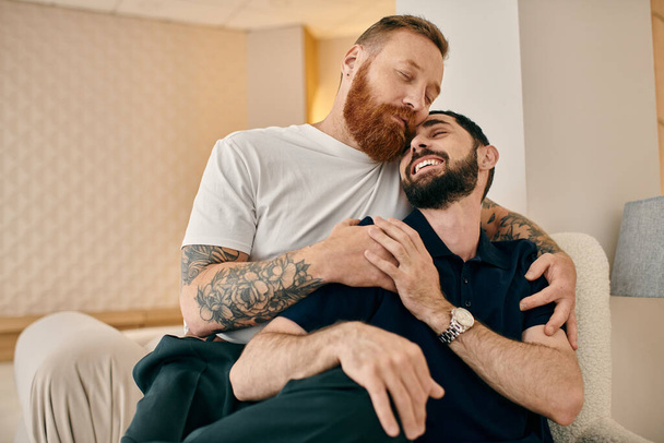 Zwei Männer in lässiger Kleidung umarmen sich herzlich auf einer gemütlichen Couch in einem modernen Wohnzimmer und drücken ihre Zuneigung füreinander aus. - Foto, Bild