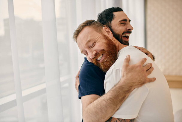 Dois homens de roupas casuais se abraçam com amor em uma moderna sala de estar, capturados em um momento terno pela janela. - Foto, Imagem