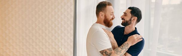 Deux hommes gays heureux en vêtements décontractés partageant un câlin chaleureux dans un salon moderne. - Photo, image