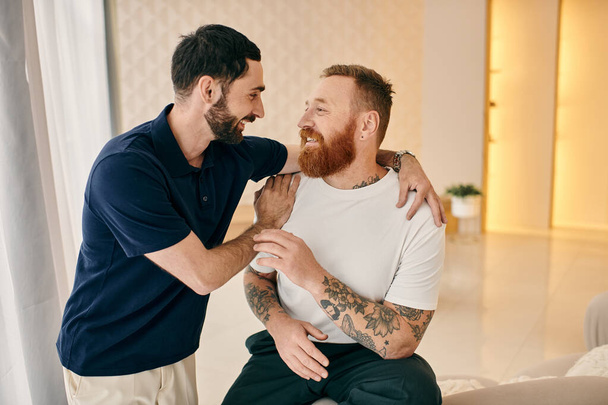 Δύο άντρες με καθημερινά ρούχα αγκαλιάζονται θερμά σε ένα μοντέρνο σαλόνι, δείχνοντας στοργή και αγάπη σε μια οικεία στιγμή.. - Φωτογραφία, εικόνα
