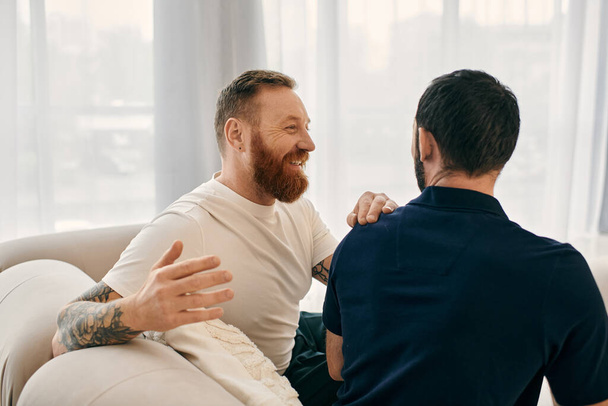 Δύο άνδρες με καθημερινά ρούχα, ένα ευτυχισμένο γκέι ζευγάρι, κάθονται σε έναν καναπέ σε ένα μοντέρνο σαλόνι, ασχολούνται με μια ζωντανή συζήτηση. - Φωτογραφία, εικόνα