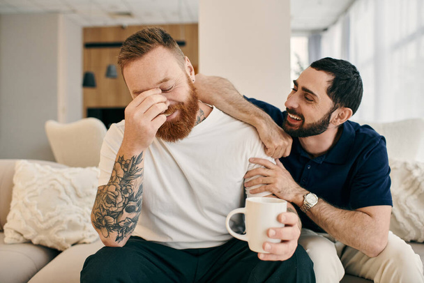 モダンなリビングルームでソファでコーヒーを楽しむカジュアルな服で幸せなゲイカップル. - 写真・画像
