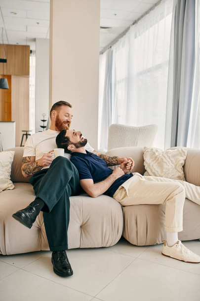 Двое мужчин, счастливая гей-пара, одетые непринужденно, сидят вместе на современном диване в гостиной, наслаждаясь компанией друг друга. - Фото, изображение