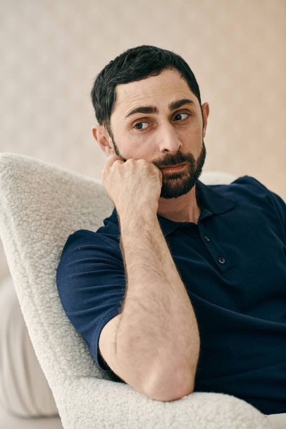 Un hombre con ropa casual, sentado en una silla, profundamente pensado con la mano en la barbilla, en una moderna sala de estar. - Foto, imagen