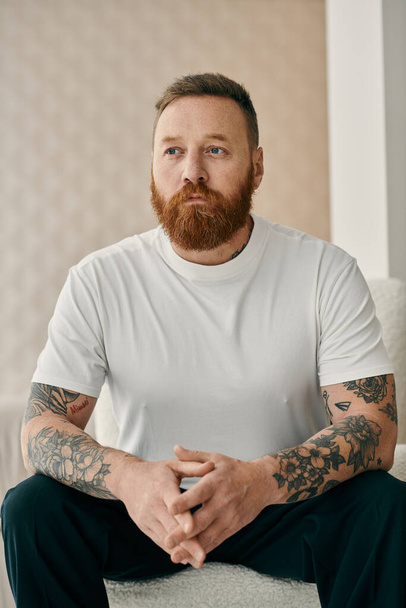Vousatý muž s tetováním sedí pohodlně na gauči, vyzařuje sebedůvěru a sebevyjádření. - Fotografie, Obrázek
