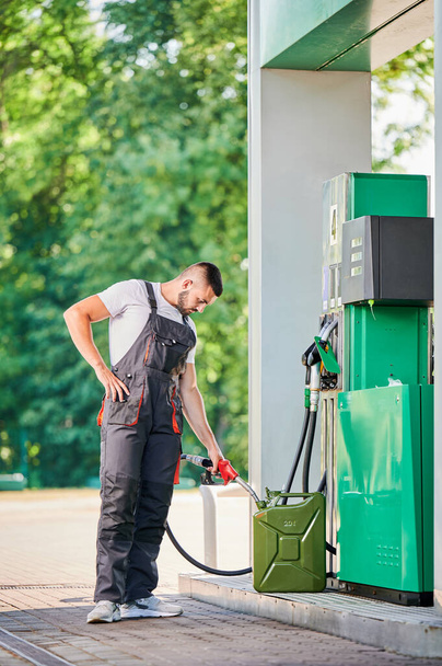 Professional worker refueling canister with fuel. Skilled man filling cistern with gasoline for customer. Man in ovealls with pump nozzle refueling canister next to gasoline column. - Photo, Image