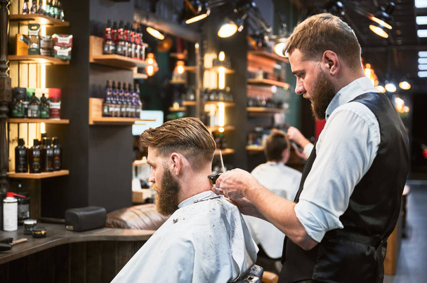 Professional barber using comb and electric shaver to cut side hair. Male customer getting stylish haircut. Side view of man on background of other hairdresser. - Photo, Image