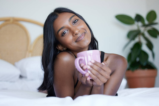 Coffee, portrait and happy woman in bedroom for morning, comfort and smile for sleep in bed. Indian female person, gen z and relax in home with drink, peace and weekend with pride in apartment. - Photo, Image