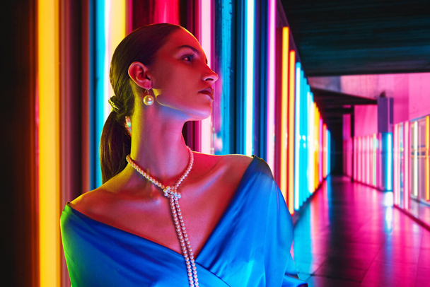 Portrait of elegant woman wearing in blue dress and pearls necklace posing looking away in neon light against striped background. Concept of youth, beauty, fashion and style, modern lifestyle. Ad - Photo, Image