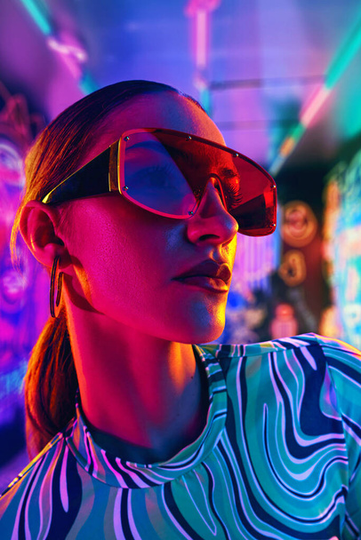 Kobieta w stylowym stroju i uderzające czerwone, ponadgabarytowe okulary przeciwsłoneczne odbijające żywe światło na neonowym tle oświetlonym graffiti. Pojęcie młodości, piękna, mody i stylu, nowoczesnego stylu życia. Ogłoszenie - Zdjęcie, obraz