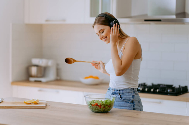 Młoda kobieta słuchająca muzyki i jedząca zdrową sałatkę z zielonymi świeżymi składnikami. głodny dziewczyna je zielony sałatka w kuchnia - Zdjęcie, obraz