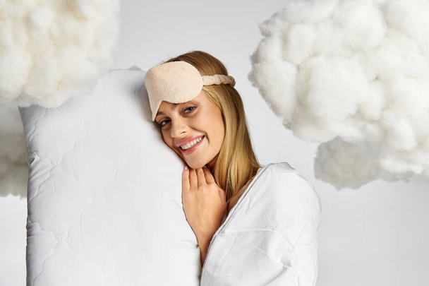A dreamy blonde woman in cozy pyjamas holds a pillow, surrounded by fluffy clouds. - Фото, изображение