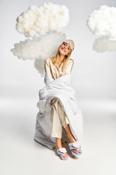 A dreamy blonde woman in cozy pajamas sits peacefully amid fluffy clouds. - Zdjęcie, obraz