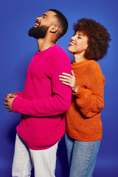Молодий афроамериканський чоловік і жінка, одягнений у яскравий повсякденний одяг, поділяють мить радості і сміху на синьому фоні. - Фото, зображення