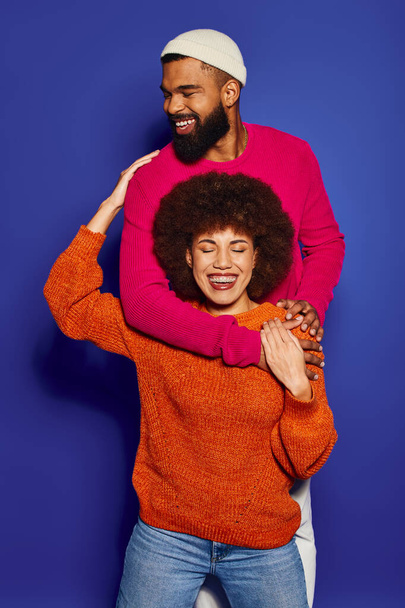 Ένας νεαρός Αφροαμερικάνος άντρας και γυναίκα, ντυμένοι με ζωντανή περιστασιακή ενδυμασία, μοιράζονται μια ζεστή αγκαλιά φιλίας σε μπλε φόντο.. - Φωτογραφία, εικόνα