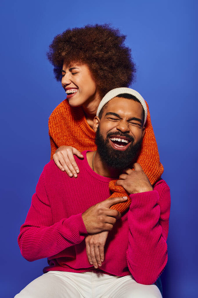 Ein Mann mit Bart umarmt zärtlich eine Frau, ihre lebendige Kleidung kontrastiert mit einem blauen Hintergrund. - Foto, Bild