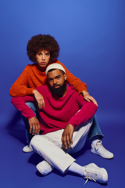 A young African American man and woman, friends in vibrant casual attire, sitting together on a blue background. - Photo, Image