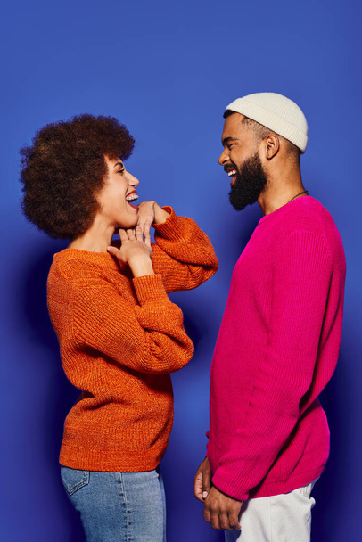 Ein junger afroamerikanischer Mann und eine junge Frau in lebhafter Freizeitkleidung teilen einen Moment des Lachens vor blauem Hintergrund. - Foto, Bild