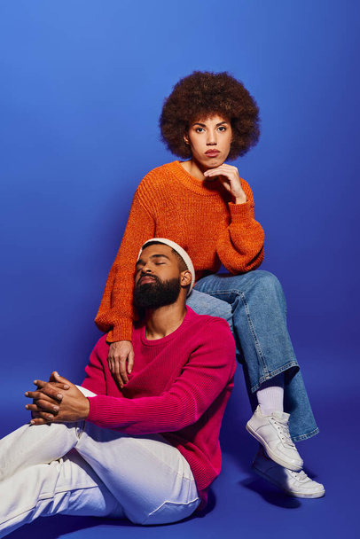 Ένας νεαρός Αφροαμερικάνος άνδρας και γυναίκα κάθονται μαζί, επιδεικνύοντας φιλία και σύνδεση, σε ένα ζωντανό μπλε φόντο. - Φωτογραφία, εικόνα
