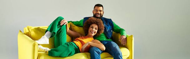Felices amigos afroamericanos con ropa vibrante sentados en un sofá amarillo sobre un fondo gris. - Foto, imagen