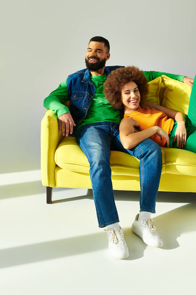 Happy African American friends in vibrant clothes, a man and a woman, sitting on a yellow couch against a grey background. - Photo, Image