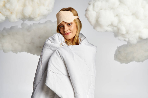 Красива блондинка, загорнута в ковдру, оточена пухнастими хмарами. - Фото, зображення