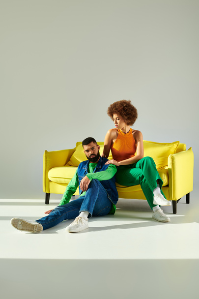 Szczęśliwy Afroamerykanin mężczyzna i kobieta w żywe ubrania salon razem na żółtej kanapie, pokazując przyjaźń. - Zdjęcie, obraz