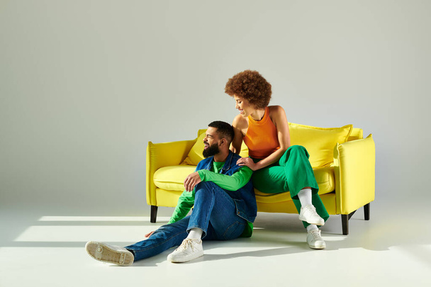 Ένας ευτυχισμένος Αφροαμερικάνος άντρας και γυναίκα με ζωντανά ρούχα κάθονται μαζί σε έναν κίτρινο καναπέ σε γκρι φόντο.. - Φωτογραφία, εικόνα