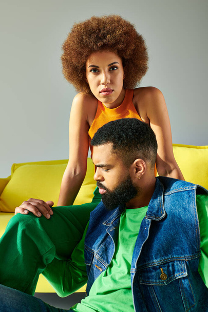 Um homem e uma mulher afro-americanos em trajes coloridos, desfrutando uns dos outros da companhia em um sofá amarelo brilhante contra um pano de fundo cinza. - Foto, Imagem