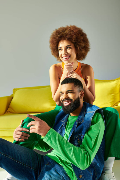 Ein glücklicher afroamerikanischer Mann und eine Frau sitzen auf einer gelben Couch und zeigen Freundschaft in lebendiger Kleidung auf grauem Hintergrund. - Foto, Bild