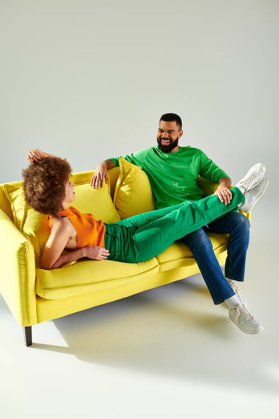 Ένας χαρούμενος Αφροαμερικάνος άντρας και γυναίκα με ζωντανά ρούχα μοιράζονται μια στιγμή φιλίας σε έναν κίτρινο καναπέ σε γκρι φόντο.. - Φωτογραφία, εικόνα