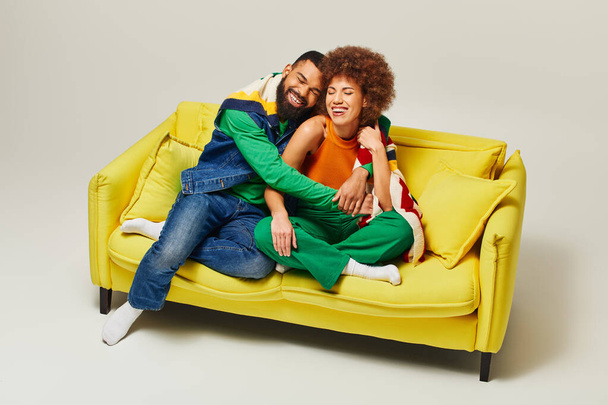 Ένας άντρας και μια γυναίκα, ντυμένοι με πολύχρωμη ενδυμασία, κάθονται ευτυχισμένοι μαζί σε έναν κίτρινο καναπέ σε γκρι φόντο.. - Φωτογραφία, εικόνα
