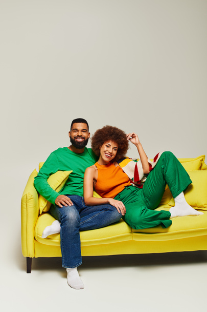 Felices amigos afroamericanos con ropa vibrante sentados en un sofá amarillo sobre un fondo gris. - Foto, Imagen
