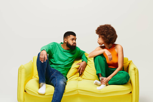 Счастливые афроамериканские друзья в яркой одежде сидят на желтом диване, демонстрируя дружбу между мужчиной и женщиной. - Фото, изображение