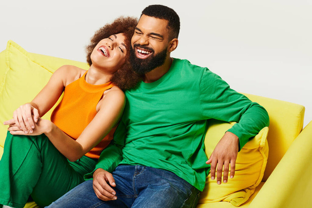 Heureux amis afro-américains en vêtements vibrants assis sur un canapé jaune sur un fond gris, mettant en valeur l'amitié entre un homme et une femme. - Photo, image