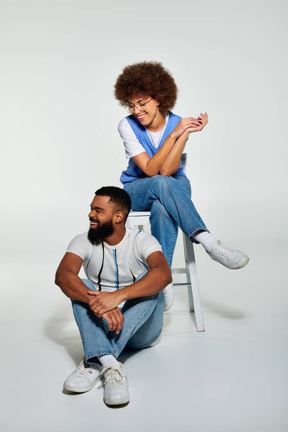 Um homem e uma mulher afro-americanos, vestidos com roupas elegantes, sentados juntos em uma cadeira contra um fundo cinza, envolvidos em uma conversa. - Foto, Imagem