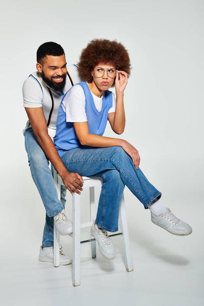 Un hombre afroamericano y una mujer, vestidos con estilo, compartiendo un taburete en una pose de amistad sobre un fondo gris. - Foto, Imagen