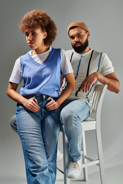 Afroameričan muž a žena ve stylovém oblečení sedí na židli, ukazuje přátelství na šedém pozadí. - Fotografie, Obrázek