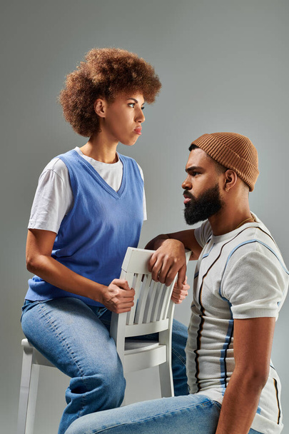 Ein afroamerikanischer Mann und eine afroamerikanische Frau in modischer Kleidung sitzen nebeneinander auf einem Stuhl vor grauem Hintergrund.. - Foto, Bild