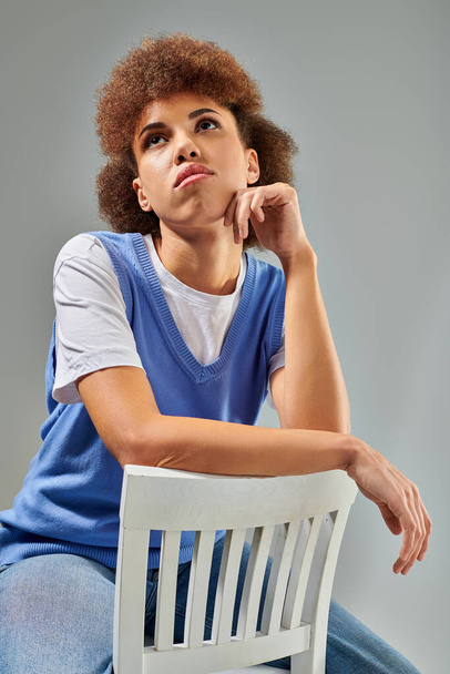 Una mujer vestida con estilo se sienta en una silla blanca, expresión reflexiva con la mano en la barbilla, sobre un fondo gris. - Foto, Imagen