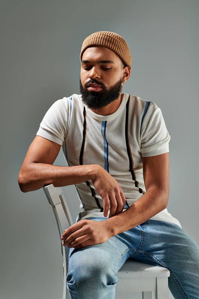 Un homme afro-américain élégamment habillé avec une barbe s'assoit élégamment sur une chaise sur un fond gris. - Photo, image
