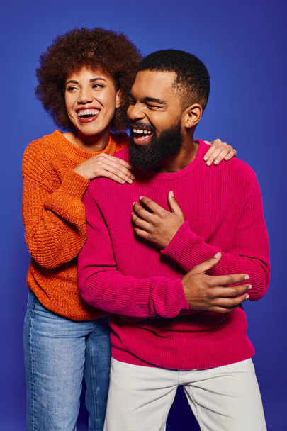 Un jeune homme et femme afro-américain, des amis, en tenue décontractée vibrante, partageant un câlin sincère sur un fond bleu. - Photo, image