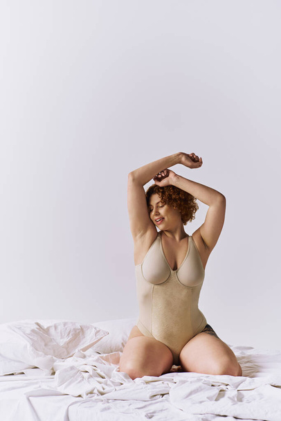 Young, curvy redhead woman in bodysuit sitting gracefully on bed against grey backdrop. - 写真・画像