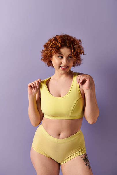 Young curvy redhead woman showcasing strength in yellow sports bra against purple backdrop. - Fotografie, Obrázek