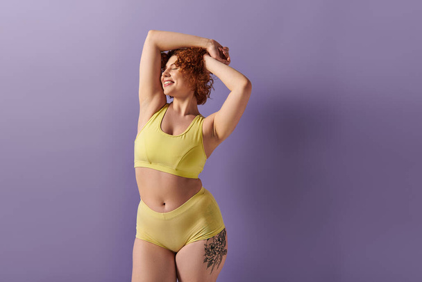A young, curvy redhead woman in a yellow bikini striking a pose on a purple background. - Foto, Imagem