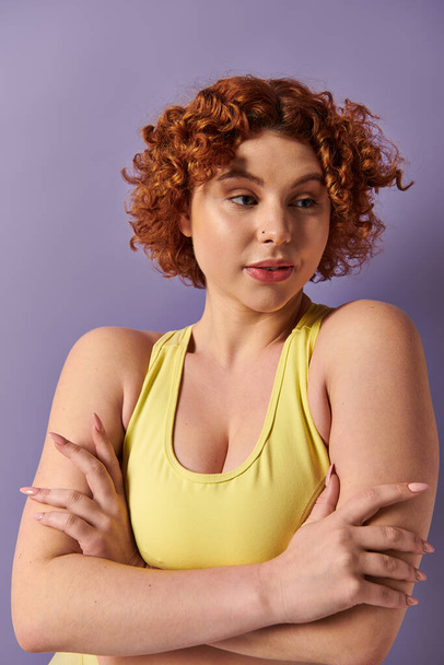 A young, curvy redhead woman in a yellow top striking a confident pose with her arms crossed. - Foto, immagini