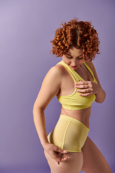 A young, curvy redhead woman in a striking yellow bikini strikes a confident pose against a vibrant purple background. - Foto, Imagem