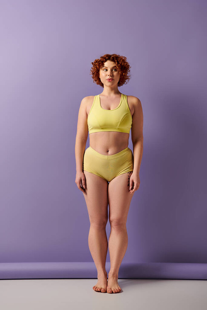 A young, curvy redhead woman in a yellow bikini stands confidently in front of a vibrant purple wall. - Foto, afbeelding