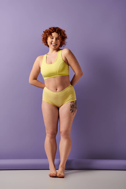 Curvy redhead woman flaunting a yellow bikini in front of a vibrant purple wall. - Photo, image